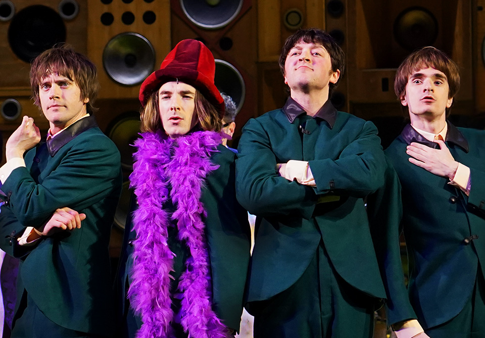 Four people in teal suits with a speaker wall background, one in a red hat and purple boa.