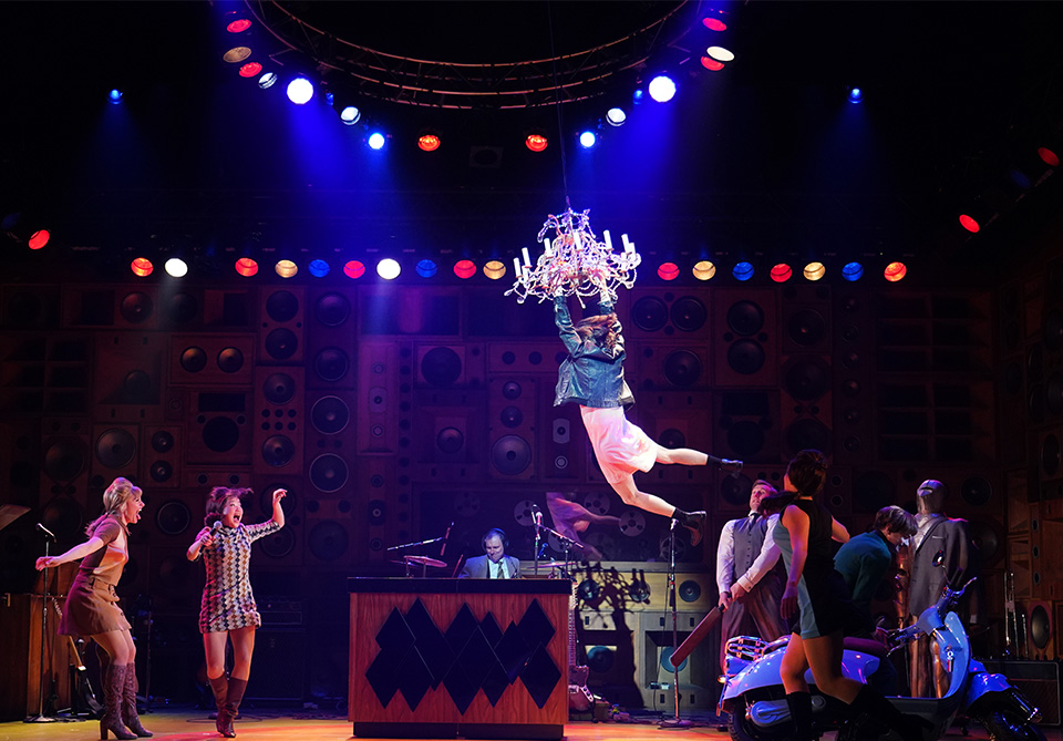 A theatrical performance with a person swinging from a chandelier on a stage with colorful lighting and a speaker-filled backdrop.