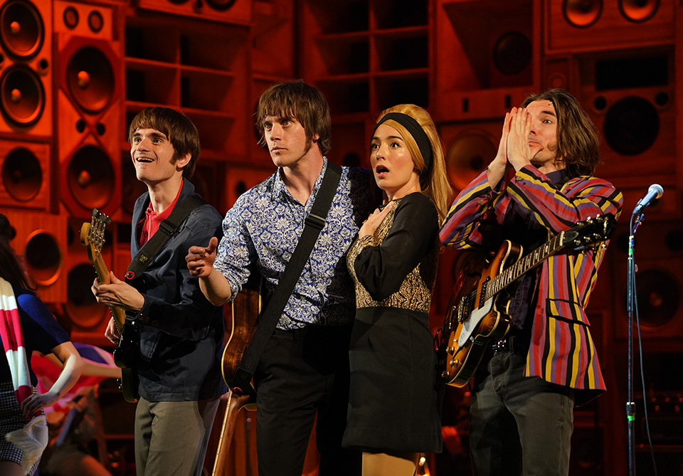 Four people on stage showing surprise, with guitars and a wall of speakers behind them.