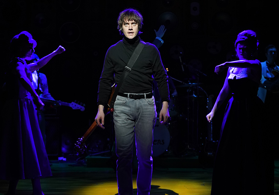 A male performer in a black turtleneck stands in a spotlight on stage, surrounded by dimly lit figures.