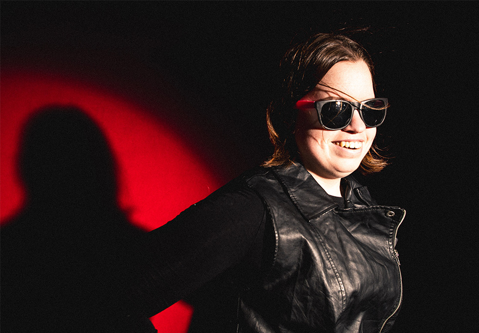 Person smiling in sunglasses and black leather jacket against a red and black background.