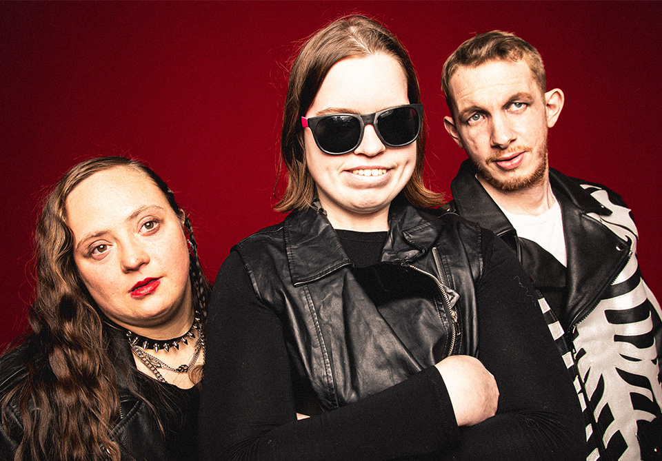 Three people in leather outfits against a red background.