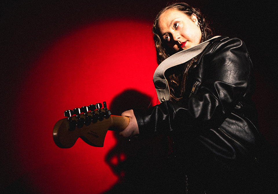 Person in a black leather jacket plays an electric guitar against a red background.