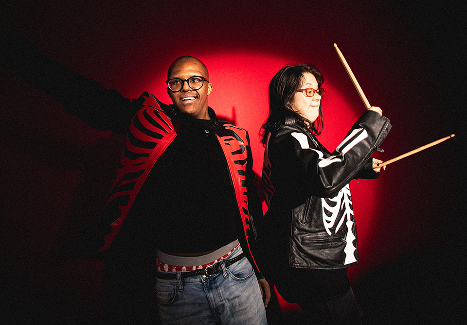 Two people wearing matching skeleton-style jackets, one holding drumsticks.