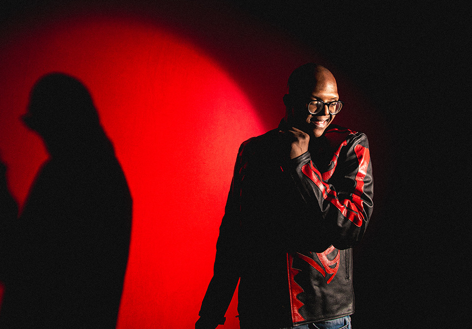 Person in a black jacket with red patterns against a red background with dramatic lighting and shadow.