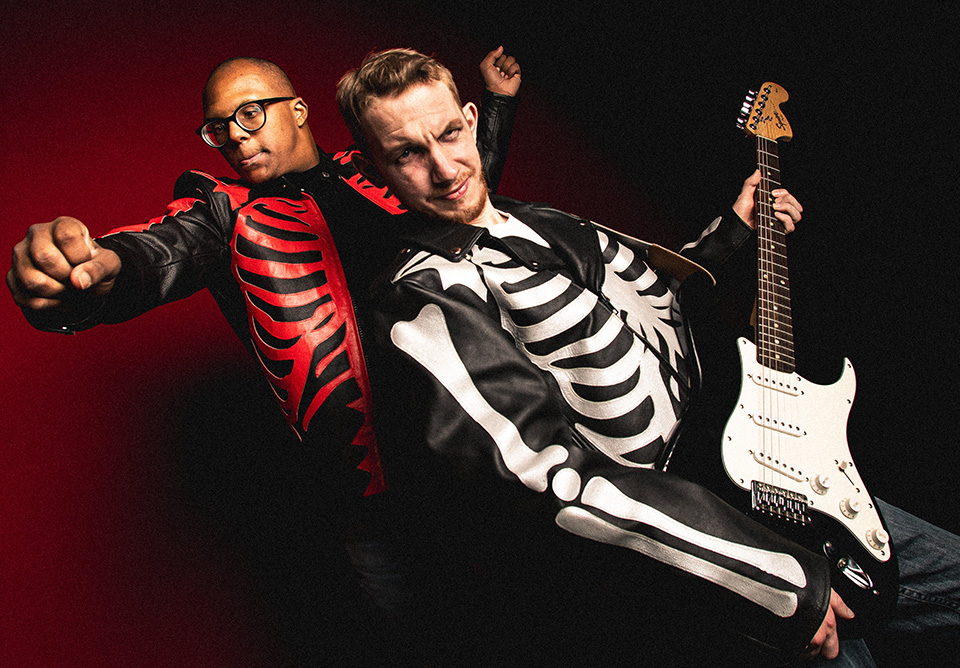 Two men in skeletal jackets pose energetically, one holding a guitar.