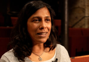 A woman with long dark hair sits in a dimly lit theater, wearing a gray top and necklace.