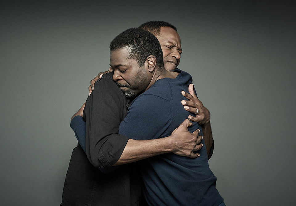 Two men embracing with eyes closed against a gray background.