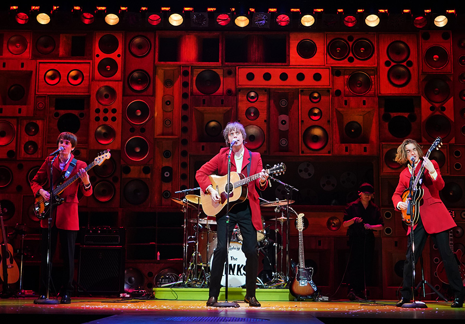 Musicians in red jackets perform on stage with a backdrop of diverse red speaker boxes.