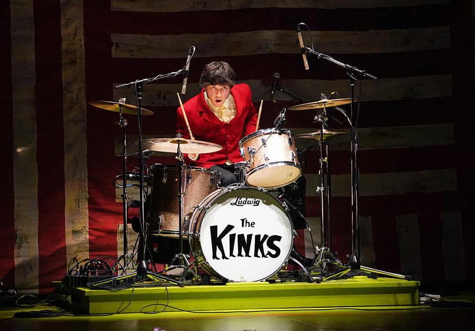 Drummer in a red blazer playing on a drum set with "The Kinks" logo.
