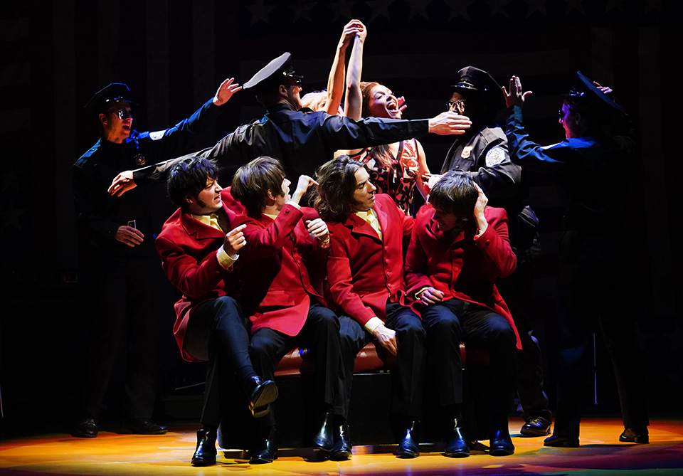 A theatrical scene with seated performers in red jackets surrounded by people in police uniforms on stage.