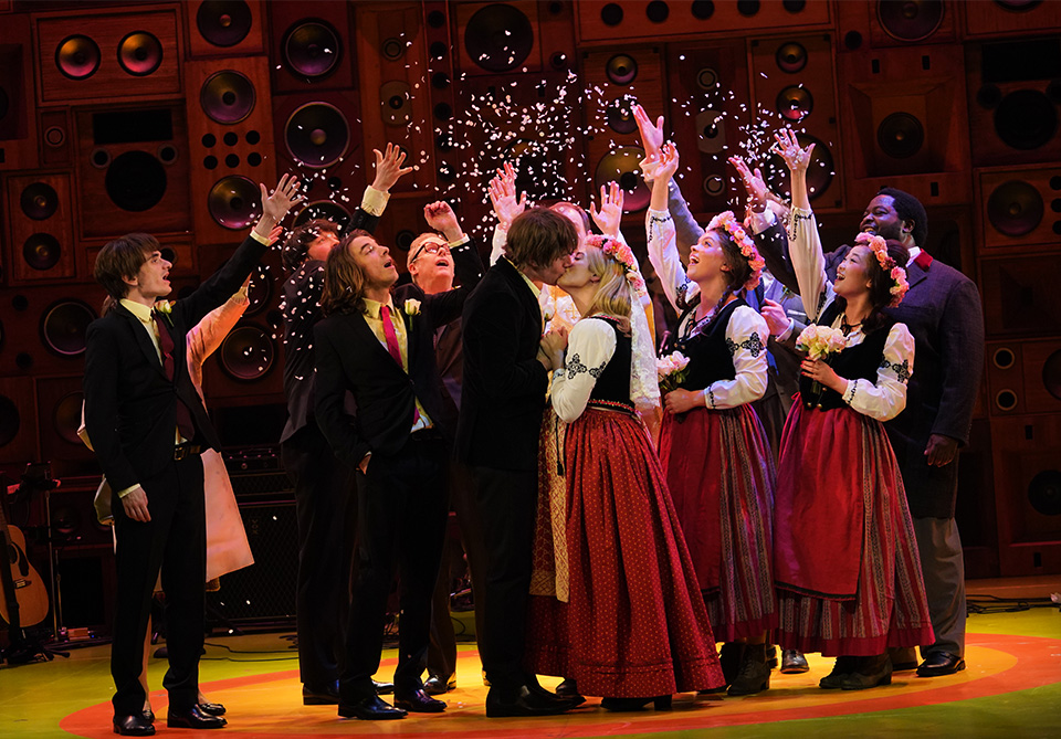 A group of people on stage in festive attire, with a couple kissing surrounded by confetti and a backdrop of speakers.