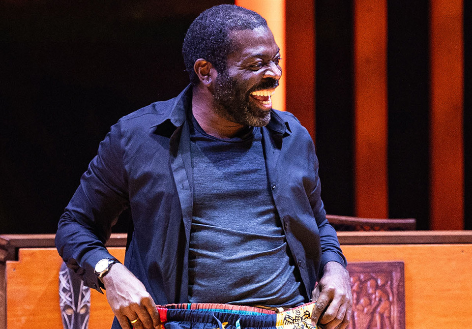 Man laughing on stage with a dark shirt and holding a multicolored patterned pair of pants.