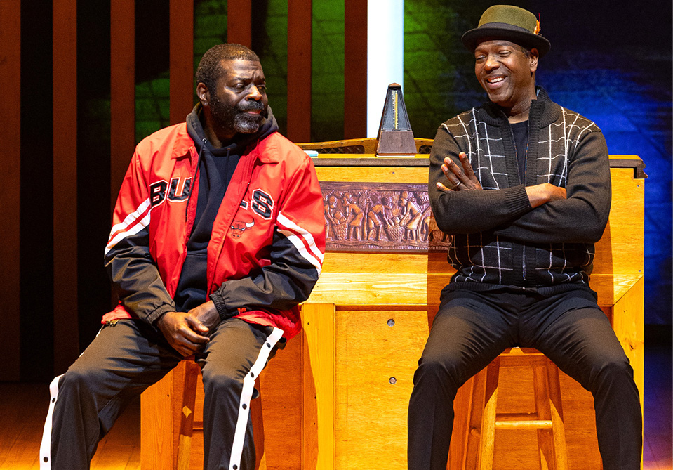 Two men sitting in front of a carved wooden piano, one wearing a red "BULLS" jacket, the other in a black hat and sweater.