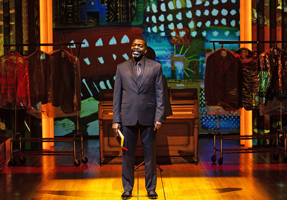 A man in a dark suit stands on a stage with colorful abstract artwork and clothing racks in the background.