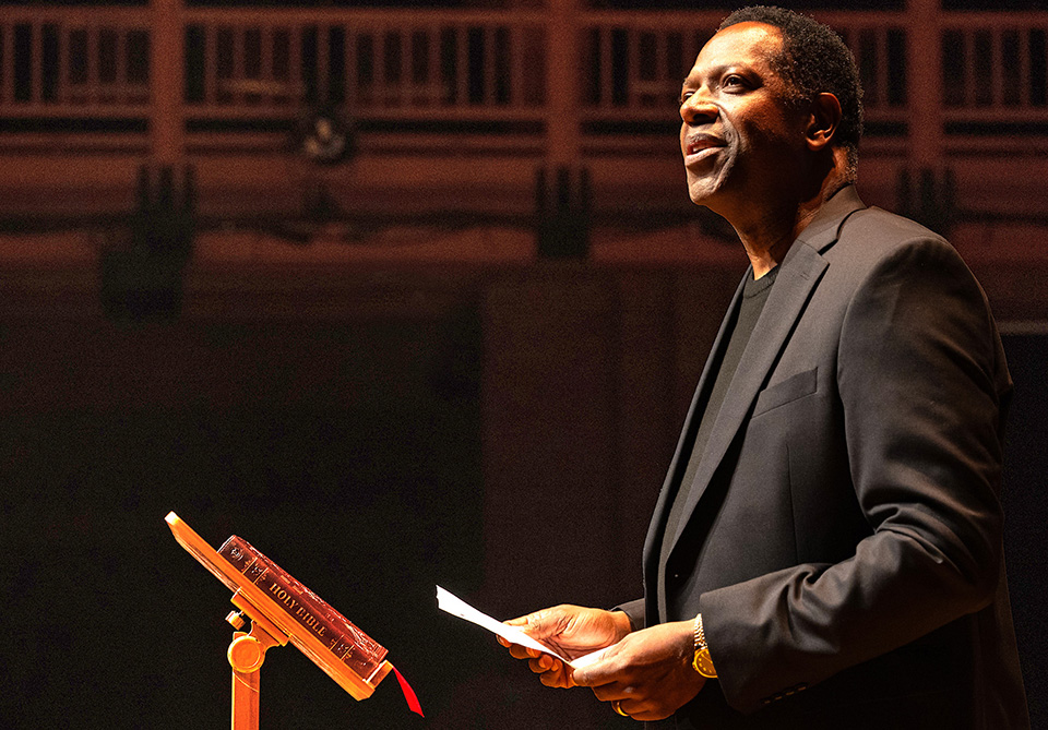 A man in a black suit speaks on stage next to a lectern with a Bible.