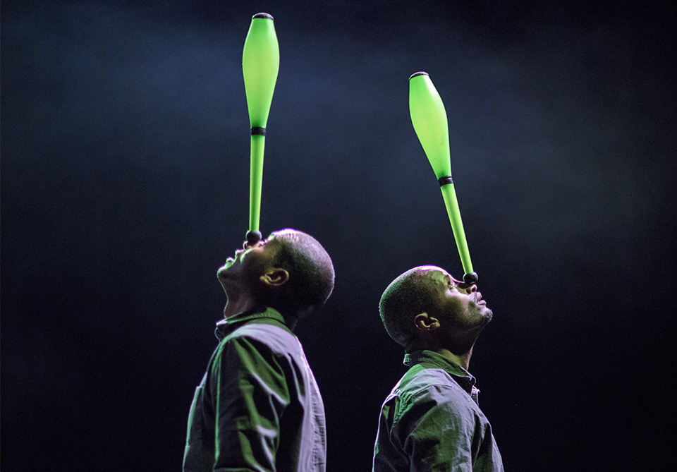 Two individuals balancing green juggling pins on their noses in a spotlight.