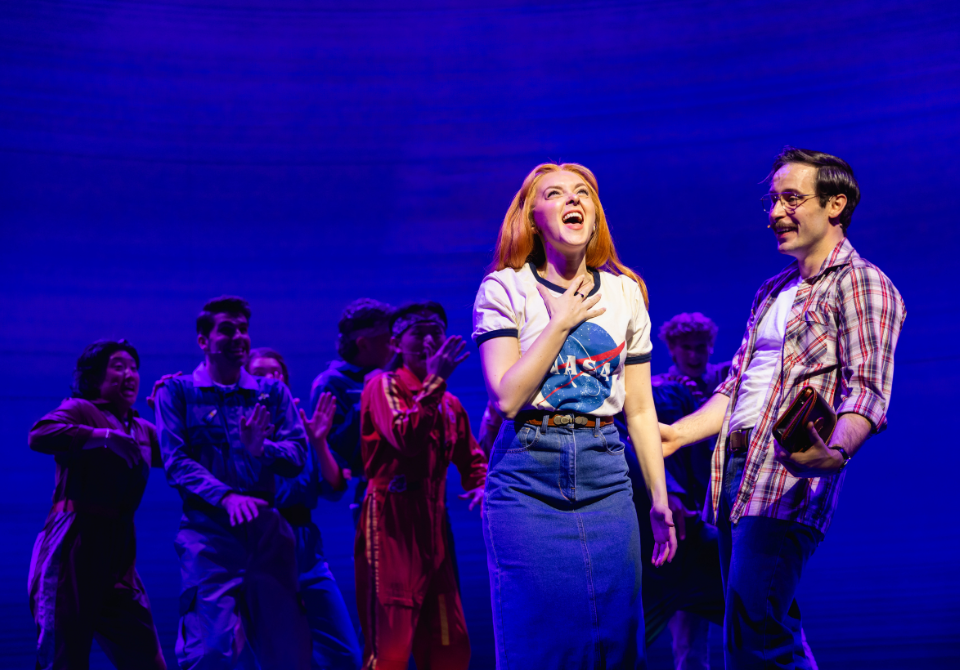 Performers on stage, a woman in a NASA T-shirt and a man in a plaid shirt under blue lighting.