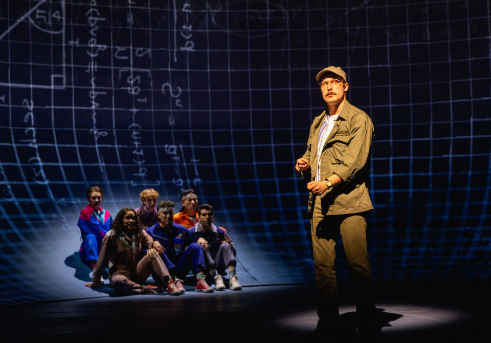 A man in a khaki jacket speaks on stage with a group sitting behind him against a mathematical grid backdrop.