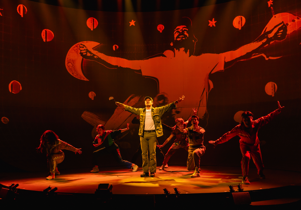 Performers on stage under red and orange lighting with a large projection background.