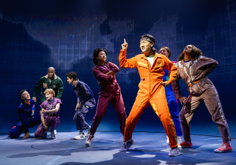 A group of performers in colorful jumpsuits on stage, with one person in an orange jumpsuit gesturing animatedly.