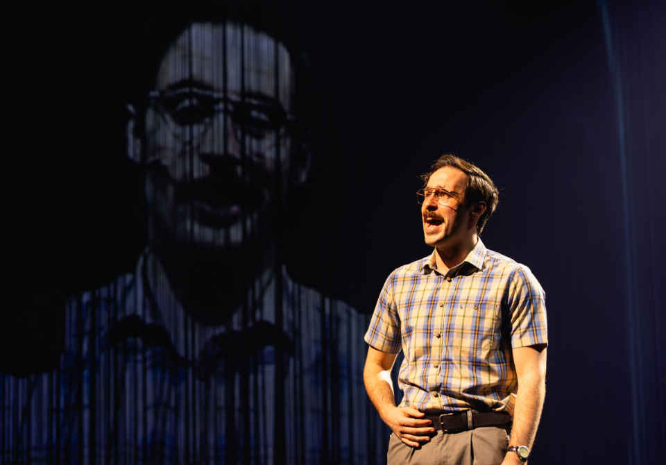 A male performer sings on stage, with a large projected image of himself in the background.