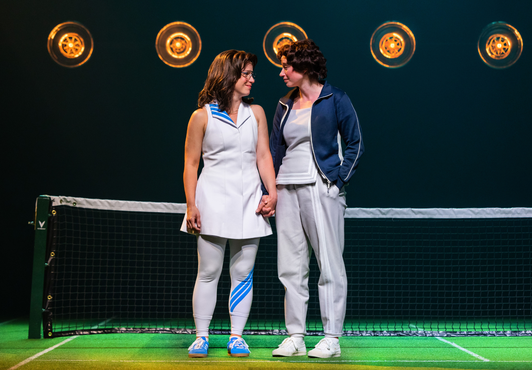 Two people holding hands on a tennis court, with a net and lights in the background.
