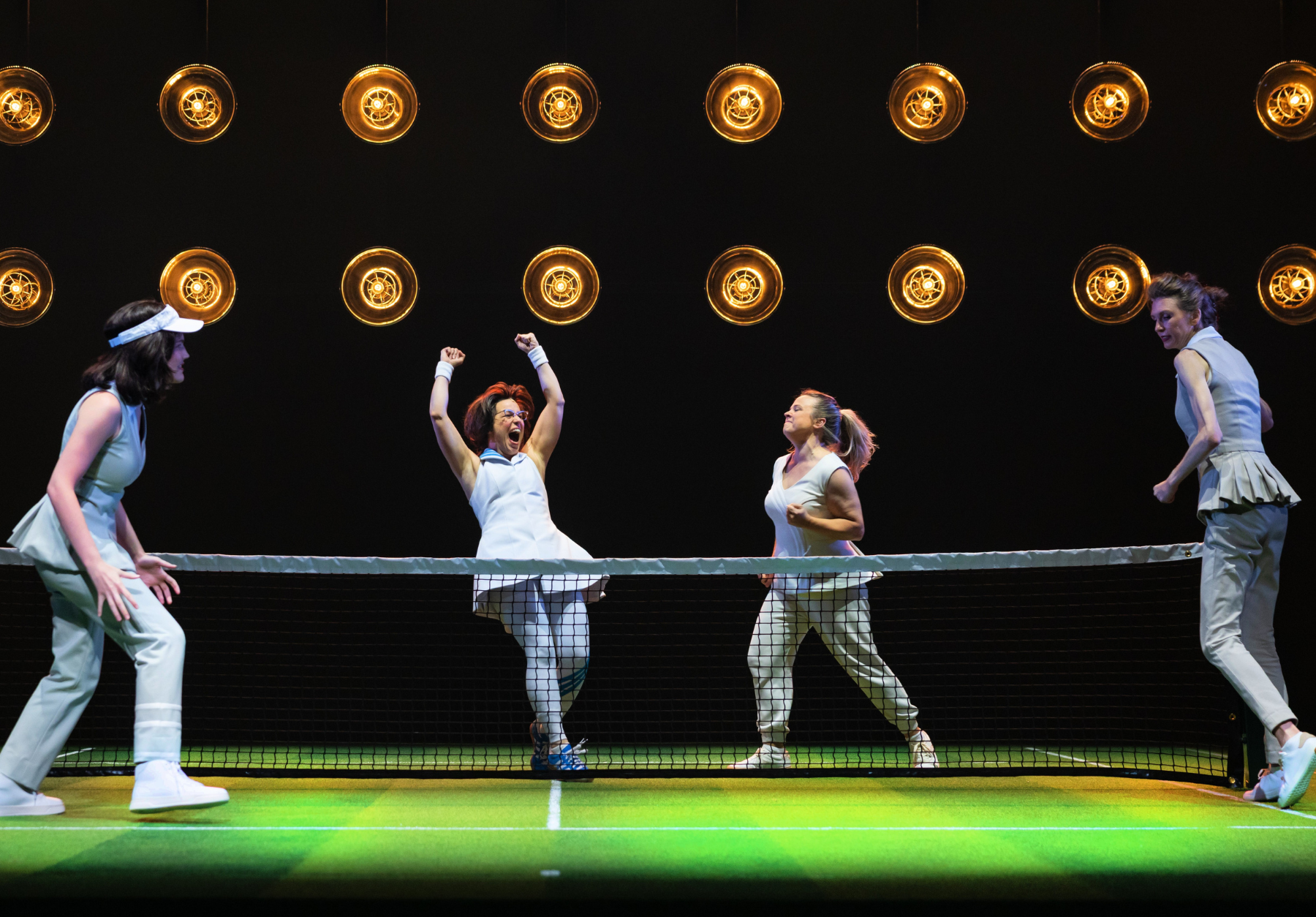 Four women in white tennis outfits perform energetically on a stage with a tennis net and overhead circular lights.