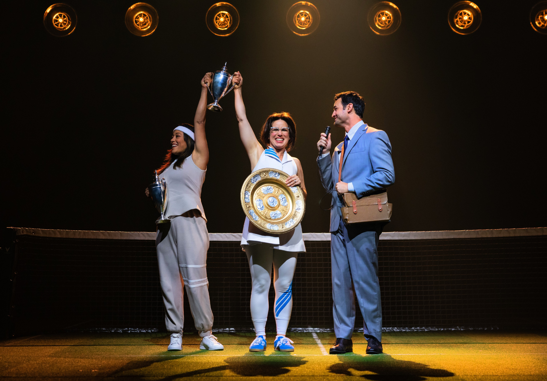Three people on a tennis court celebrating with trophies, under bright stage lights.