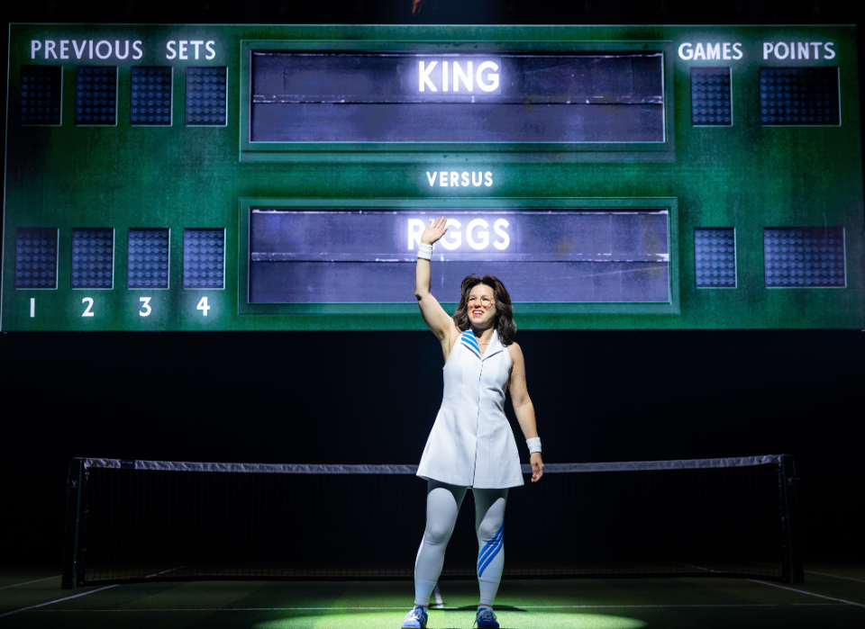Person on a tennis court waving in front of a scoreboard labeled "KING versus RIGGS."