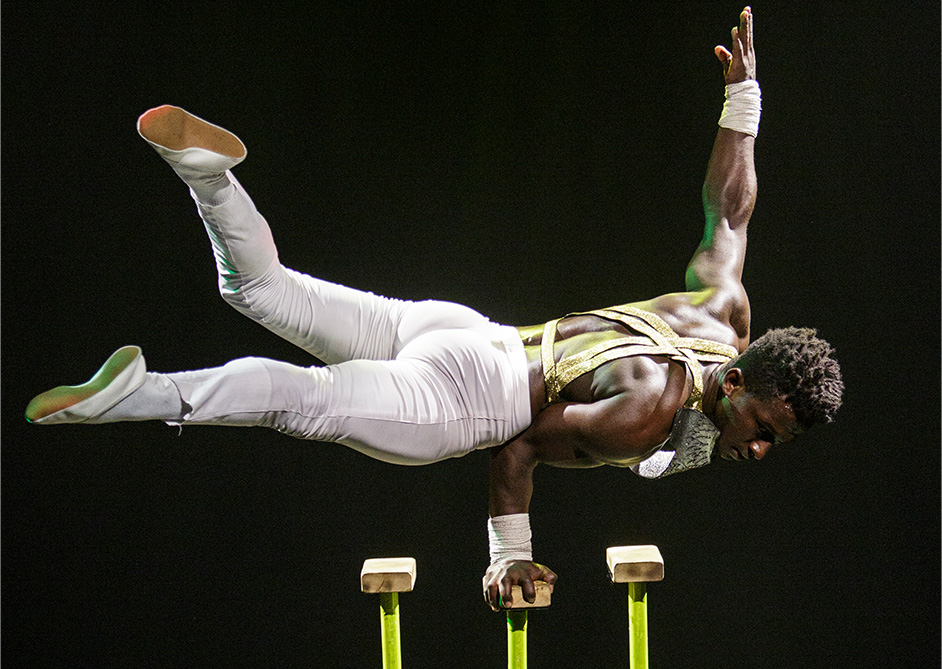 An acrobat performs a one-handed handstand on gymnastic blocks, wearing a white and gold outfit.