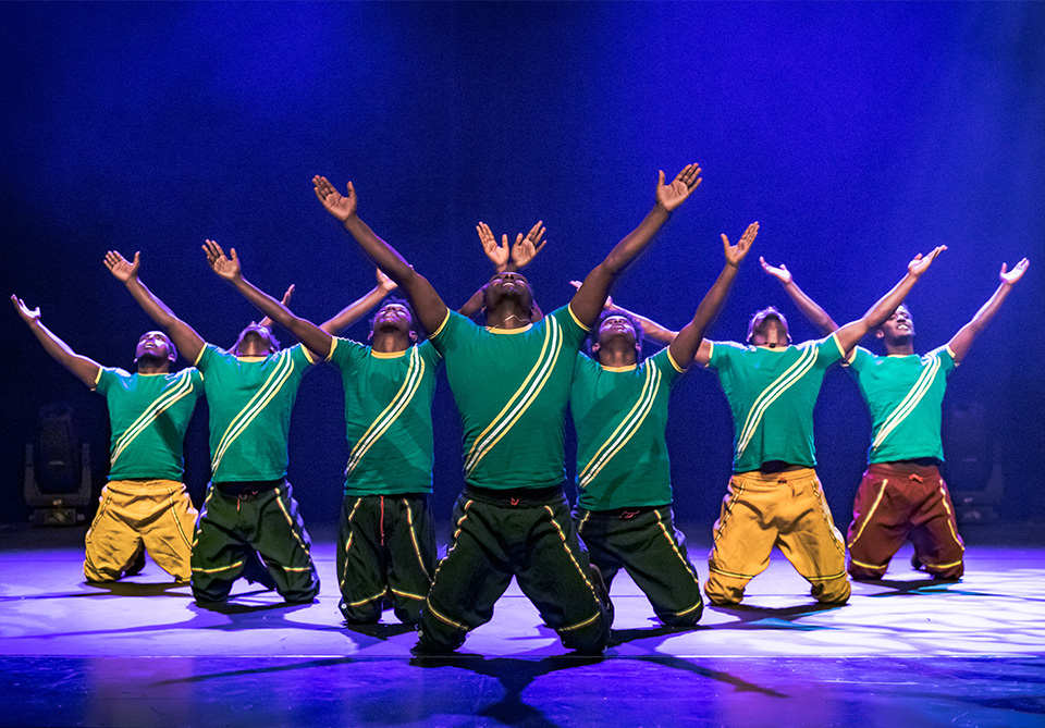 Group of performers in teal shirts with stripes, kneeling on stage with arms raised under blue lighting.