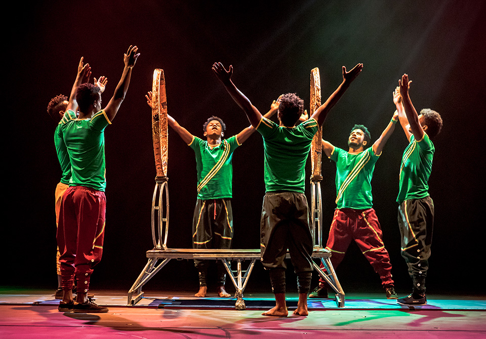 Performers in green shirts with arms raised around a metallic structure on a dimly lit stage.
