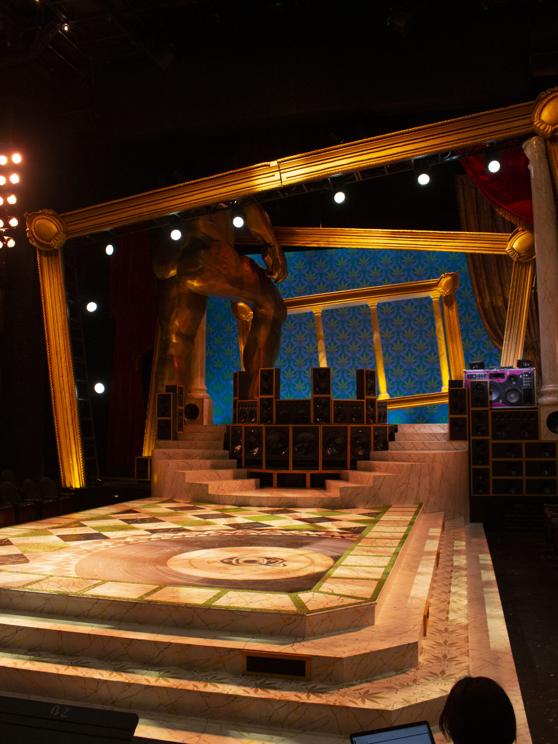 A luxurious stage set with a marble-like platform, golden statue, patterned backdrop, and stacked speakers.