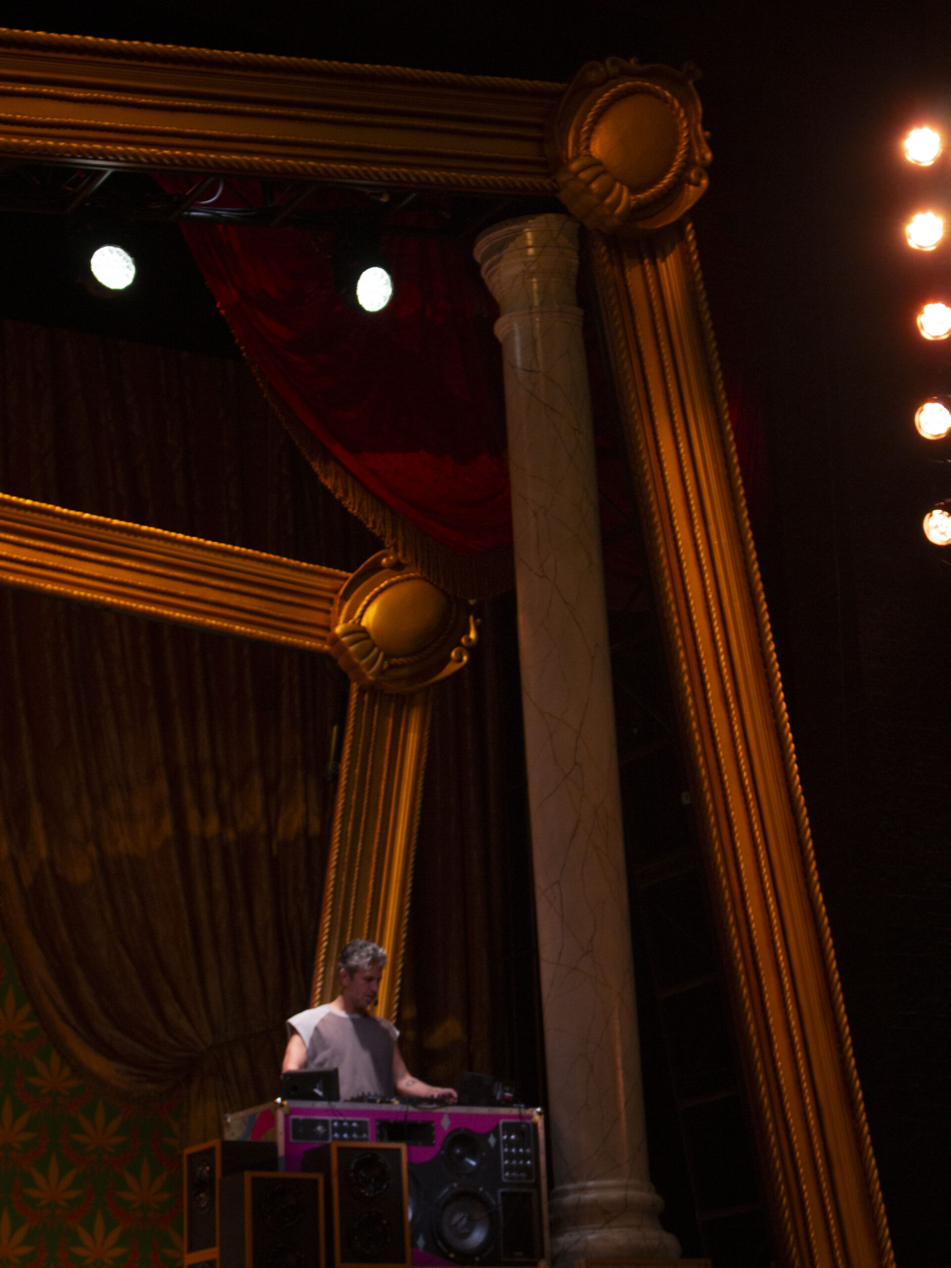 A stage with an ornate golden frame, large column, and person at a DJ console, surrounded by red curtains and pattern walls.
