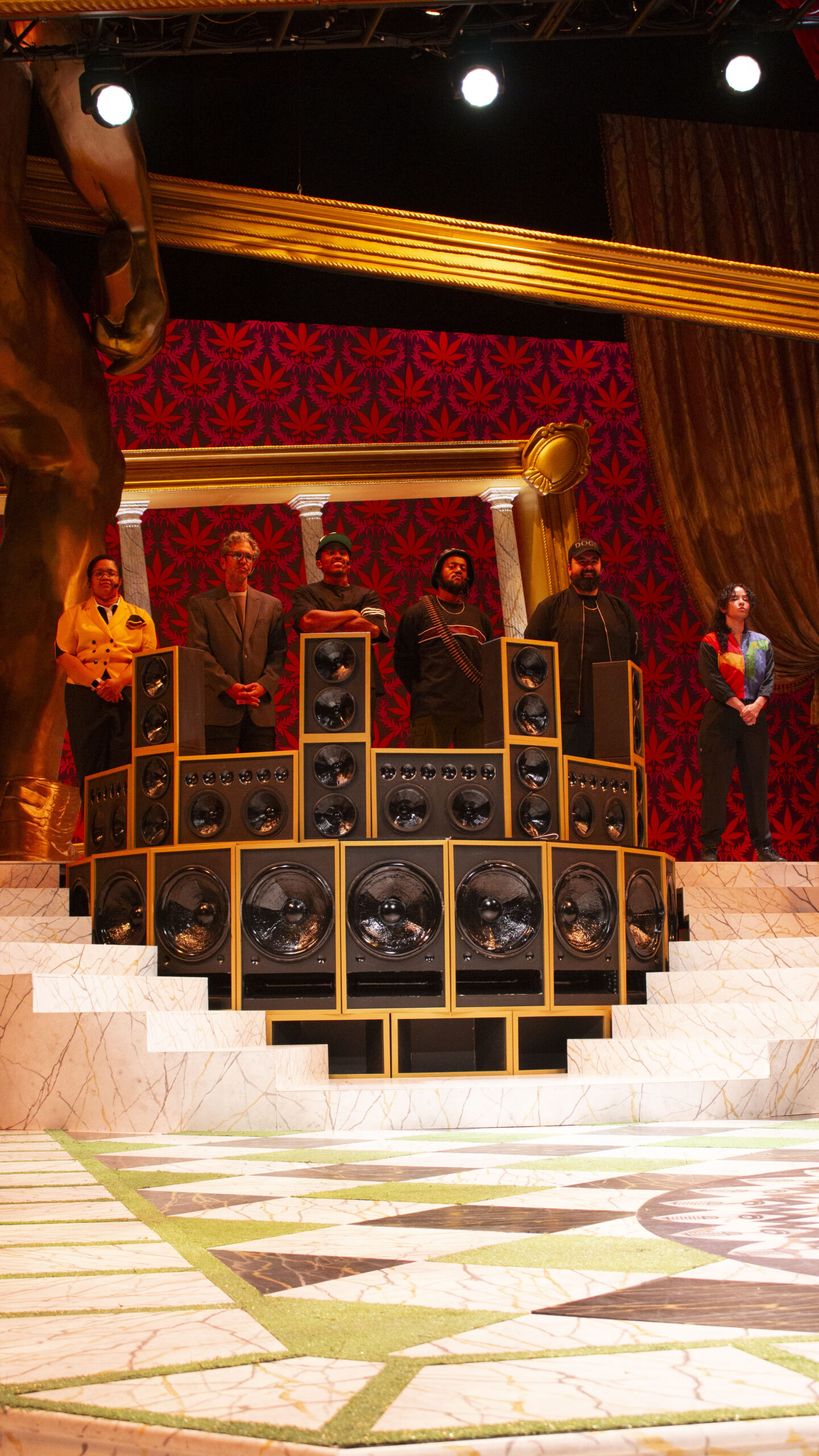 A group of people stand behind a large speaker setup on an ornate stage with a red and black backdrop and marble steps.