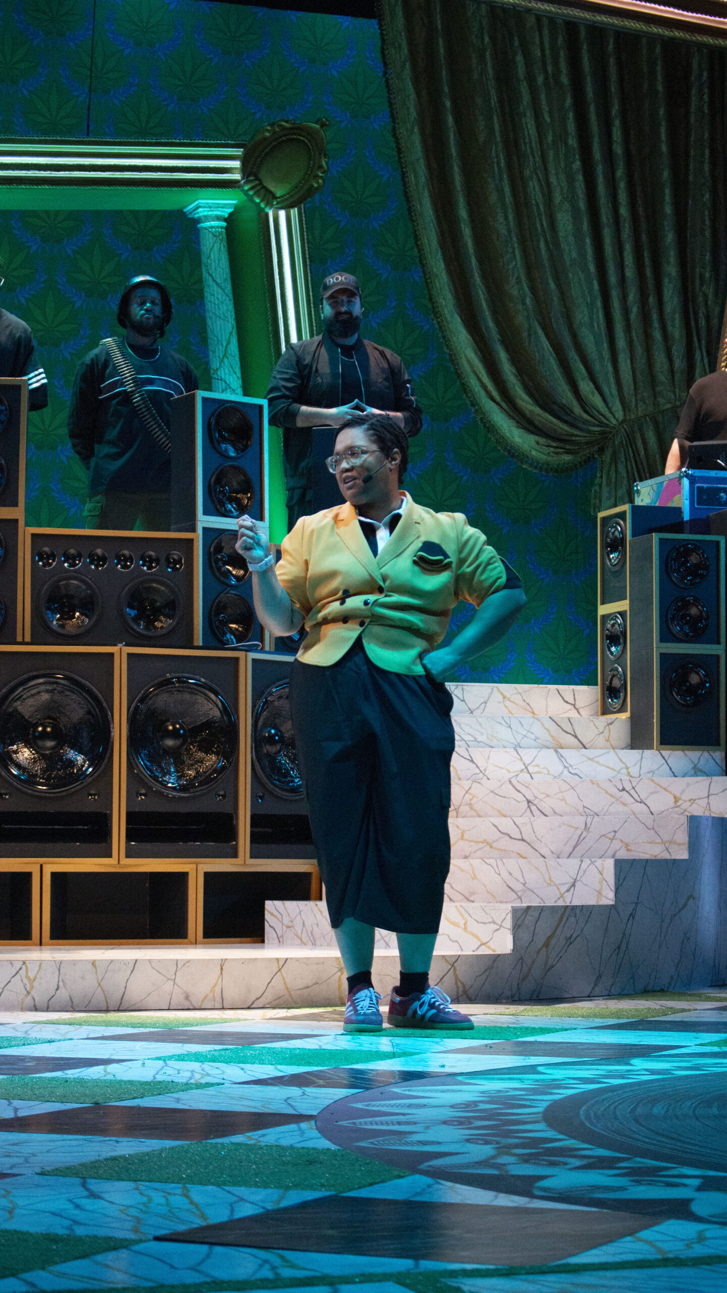 a person with a black skirt and yellow jacket on stage looking off to the side while people behind her stare out