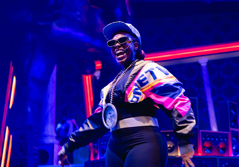 Performer in a colorful jacket and clock medallion on stage with neon lighting.