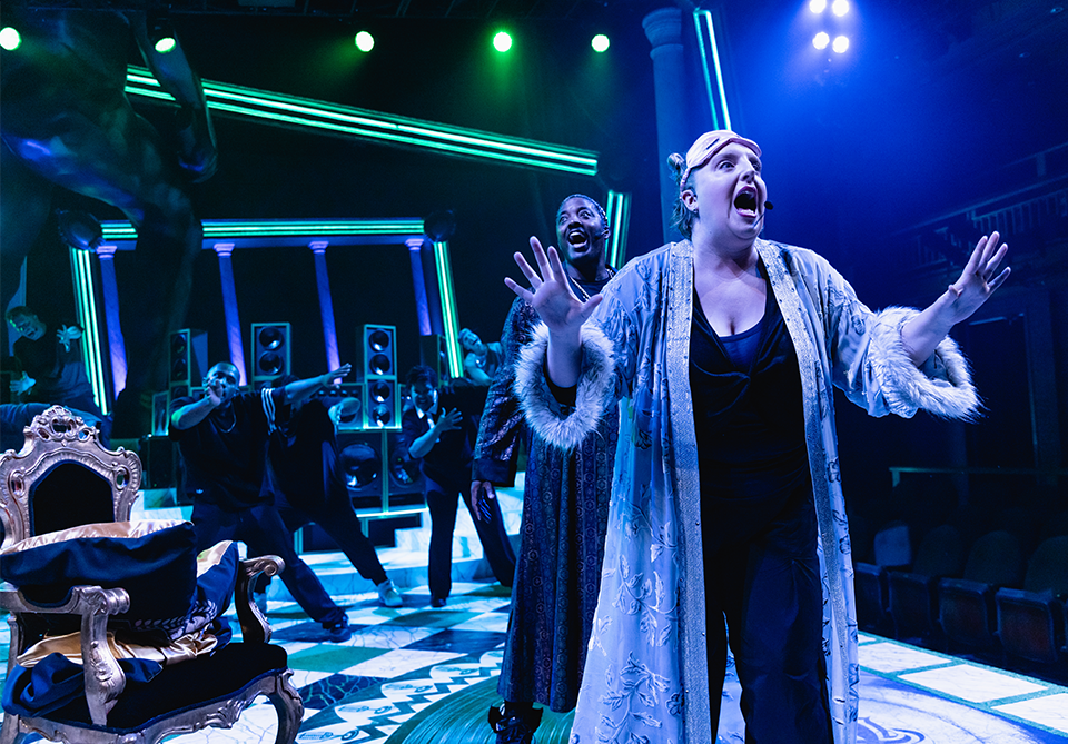 A dynamic stage performance with actors in expressive poses, vibrant lighting, and a lavish, ornate chair in the foreground.