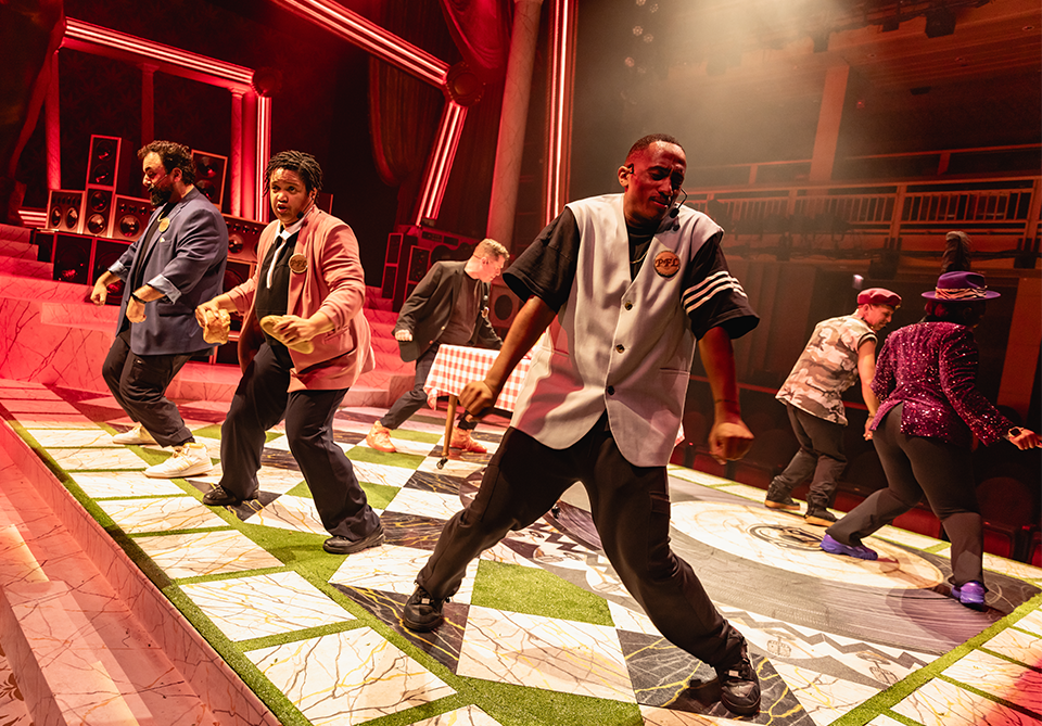 Performers dancing on a brightly lit stage with geometric floor tiles and a vibrant background.