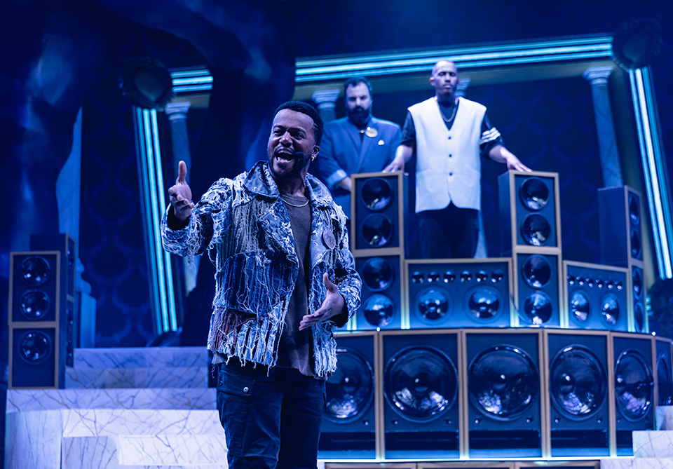 A man performs energetically on stage with large speakers and two individuals behind him, set in a blue-lit theatrical environment.