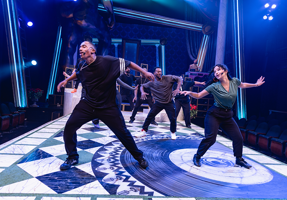 Three dancers perform energetically on a stage with a vinyl record pattern on the floor and dramatic lighting.