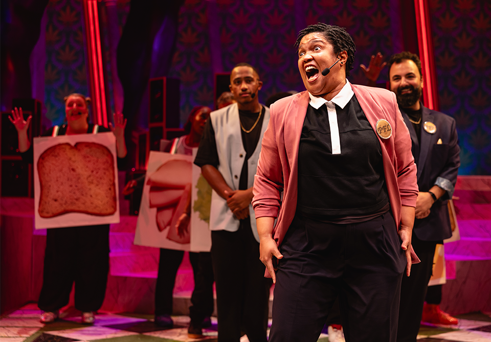 A lively scene from a theatrical performance with a person in a pink jacket speaking into a headset microphone, surrounded by performers in a colorful set.