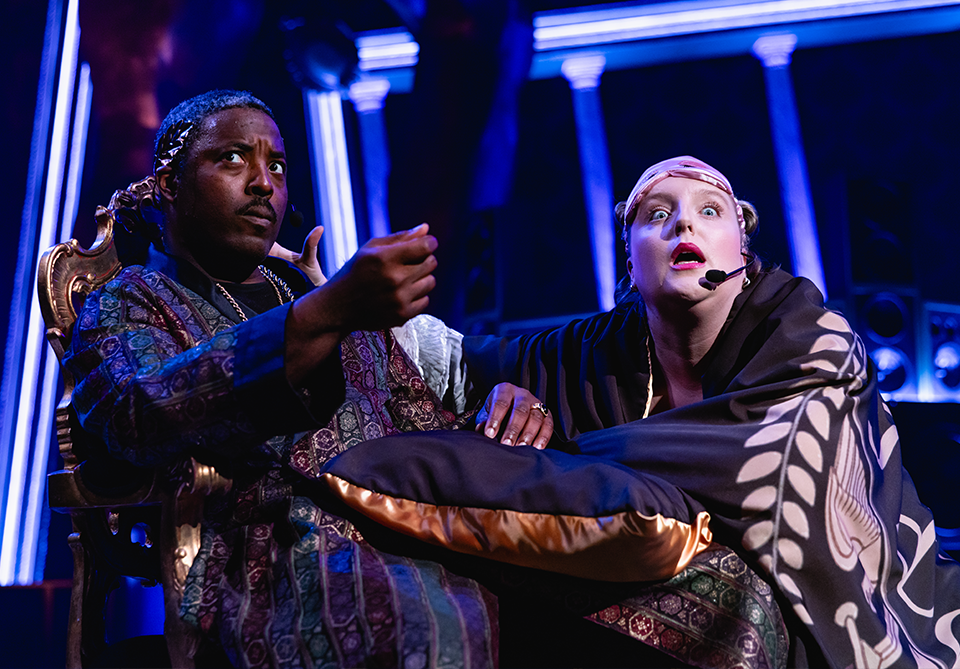 Two performers on stage, one in a multicolored robe and the other draped in a patterned shawl, both with expressive faces in a dramatic scene.