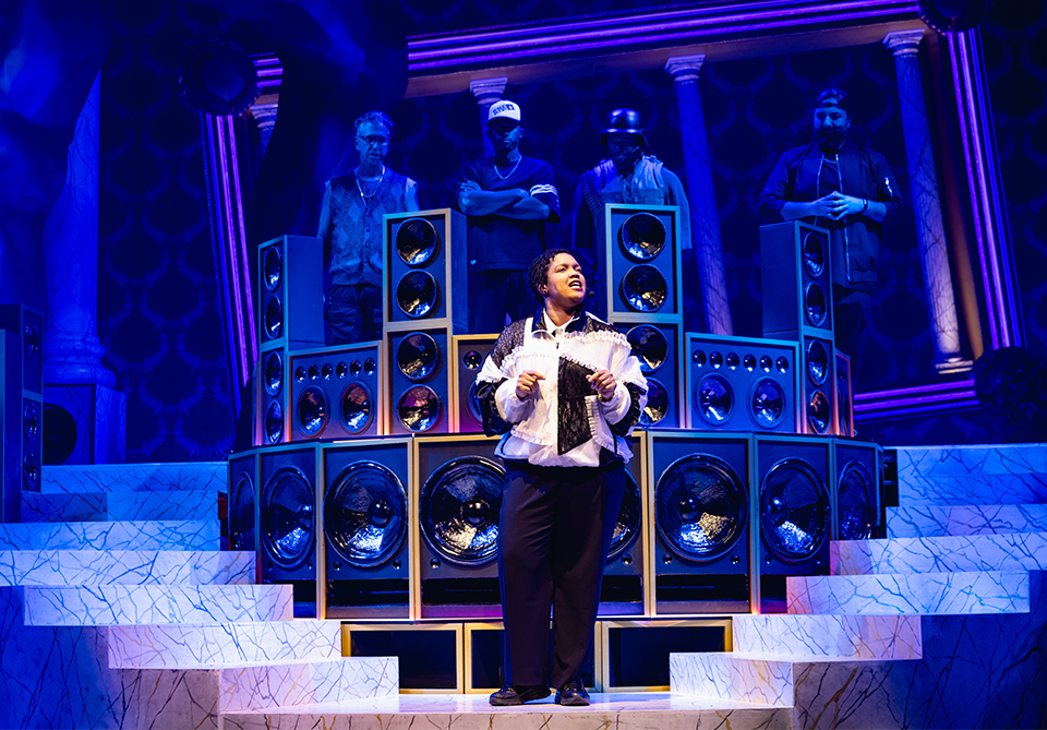 Person standing on stage surrounded by large speakers, with additional figures in the background, under blue lighting.