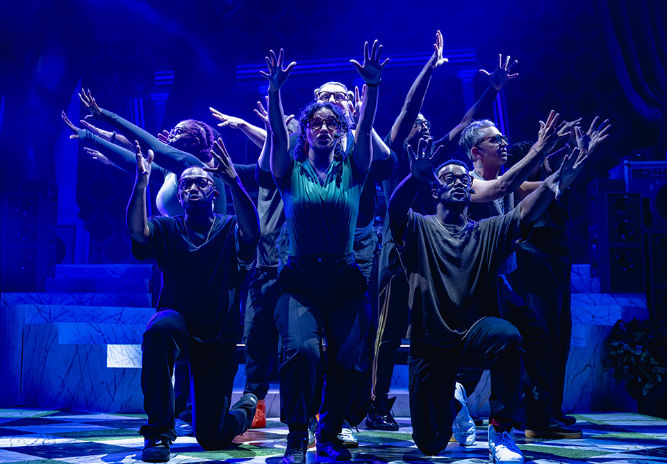 A group of performers on stage with arms raised under blue lighting.