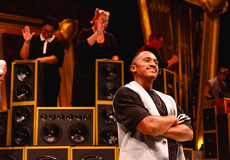 A performer stands in front of stacked speakers with arms crossed, smiling confidently on a vibrant stage.