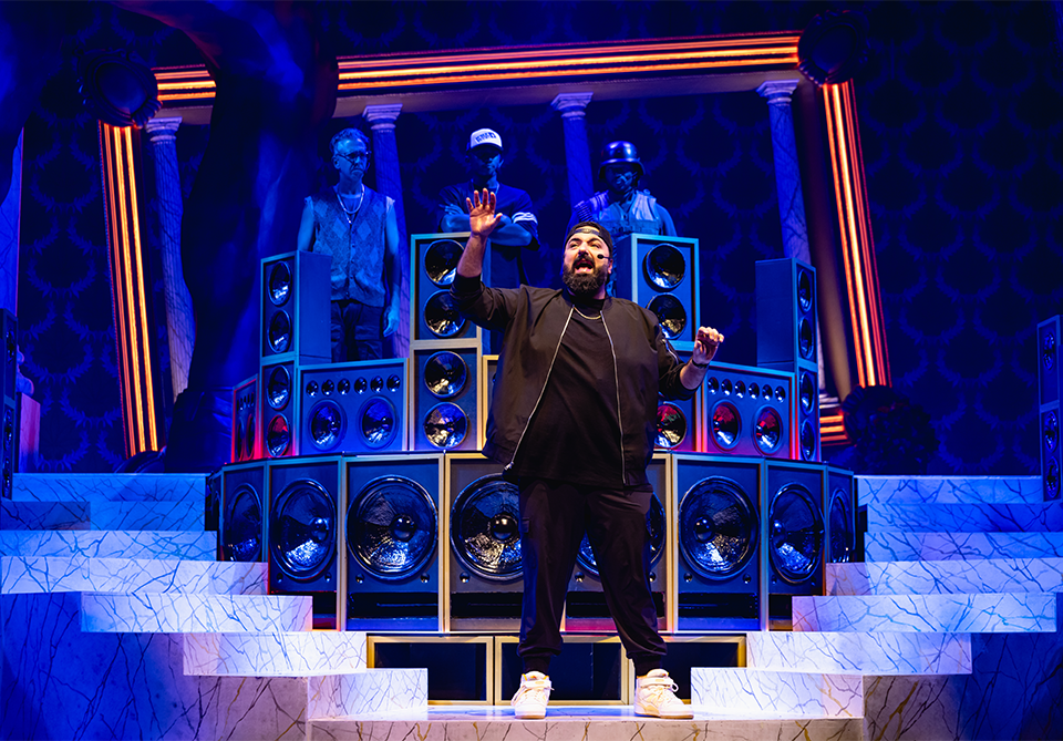 A performer stands on a marble staircase in front of large speakers and figures, under dramatic blue and orange lighting.