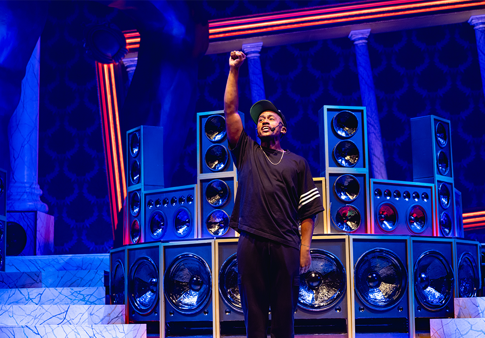 Person performing on stage with speakers and decorative pillars in the background, illuminated by blue and orange lighting.