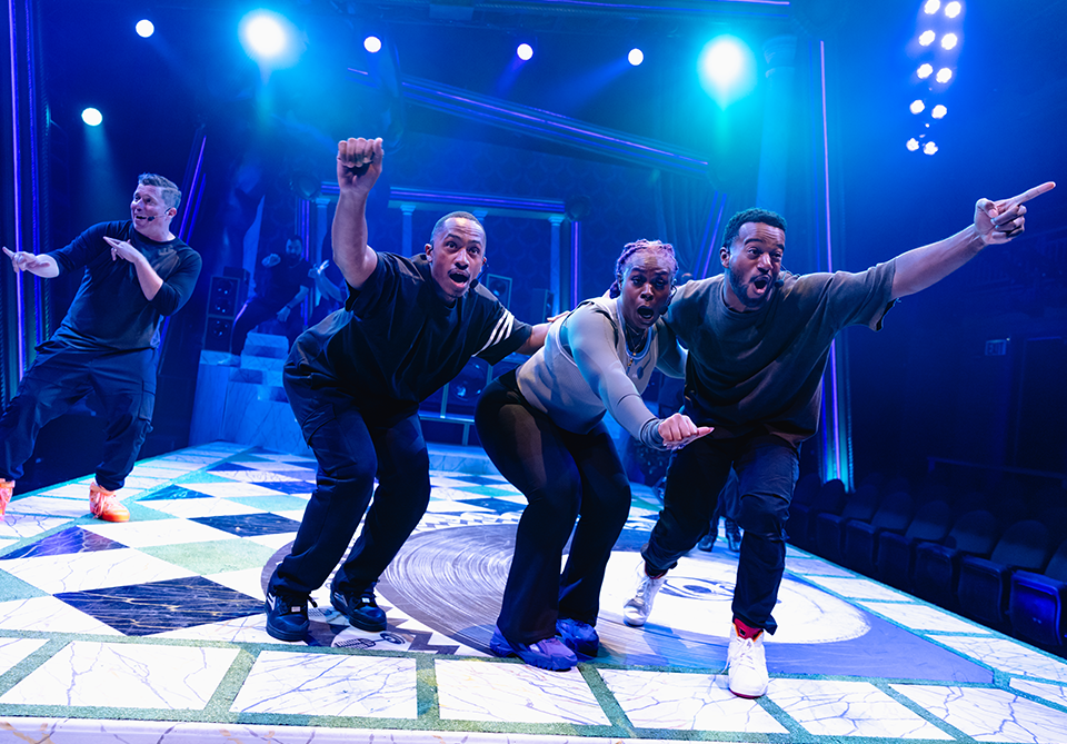 Four performers energetically pose on a brightly lit, checkered stage with colorful lighting.